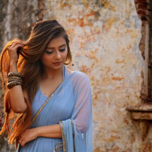 A woman in a blue saree poses gracefully outdoors by a rustic wall.