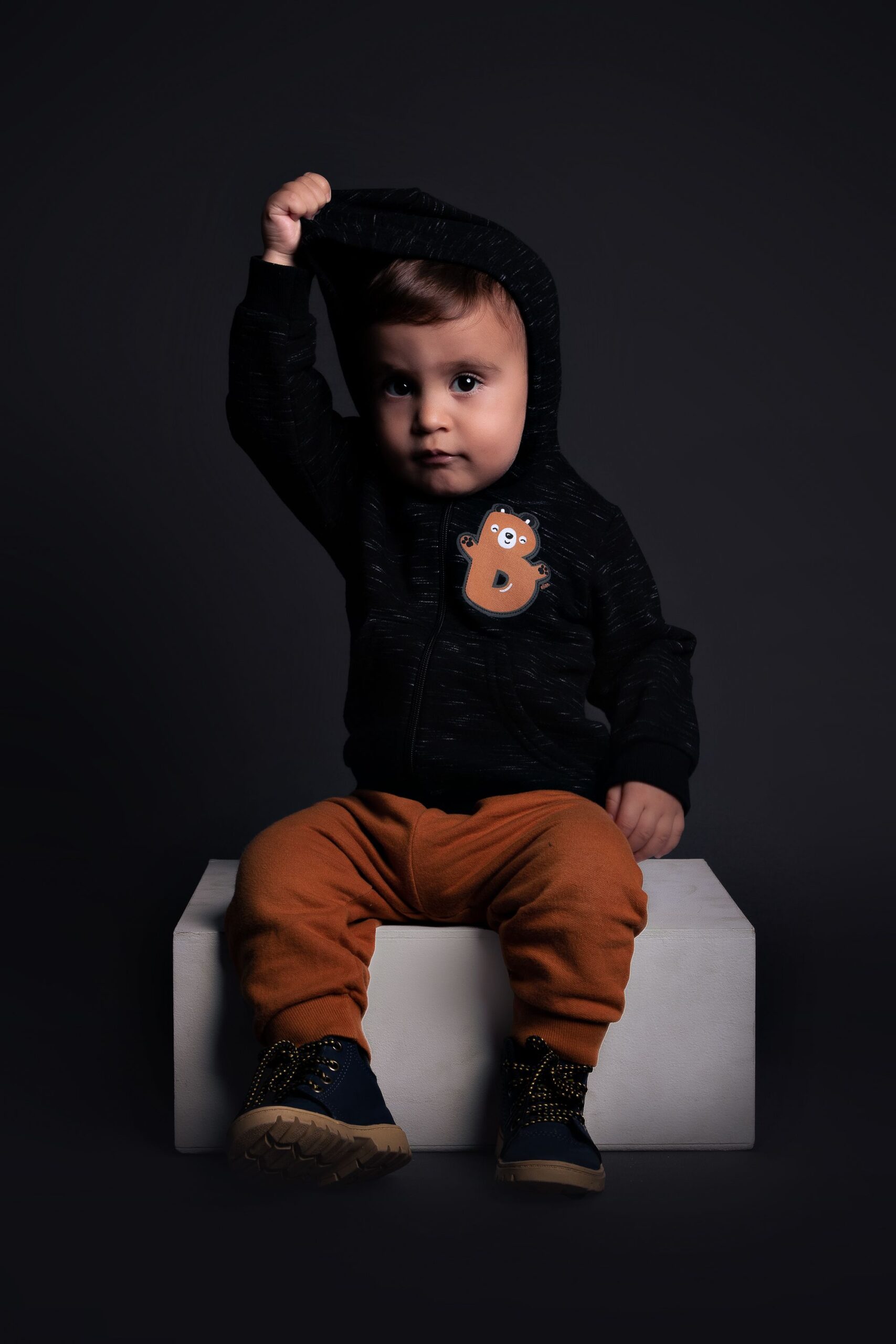 Charming portrait of a toddler wearing a stylish hoodie, posing indoors against a dark background.